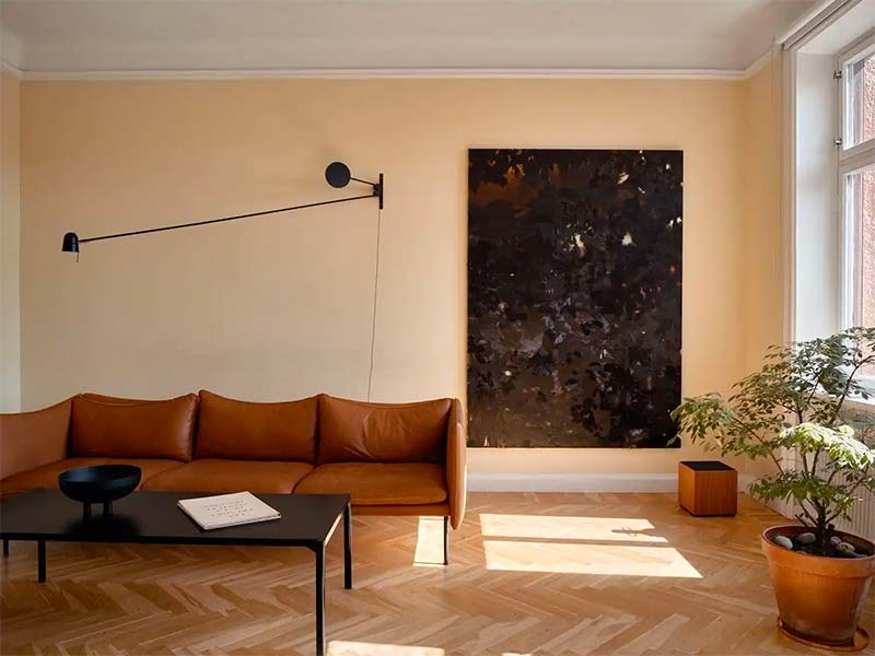 Stijlvol interieur met een visgraat houten vloer, lichtgele muren en een cognac kleur leren bank. De zwarte accenten, zoals de salontafel, de wandlamp en de grote schilderij passen er ook perfect bij.