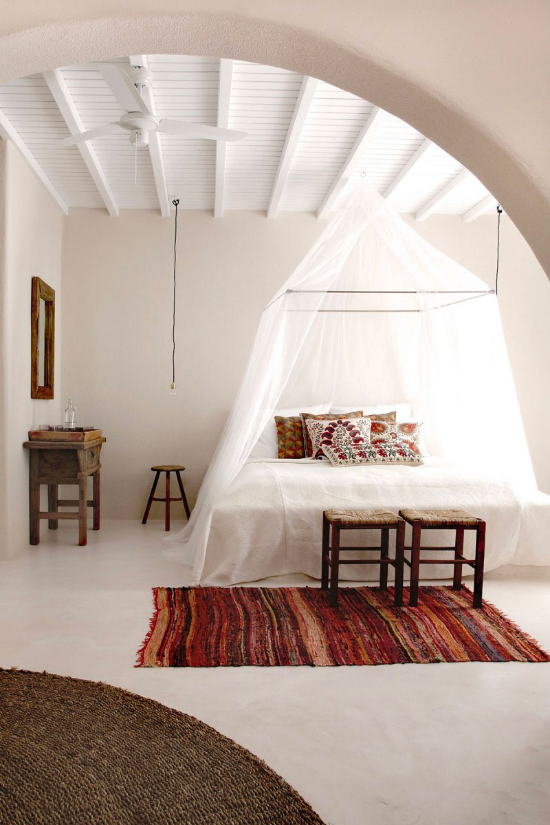 De slaapkamer van het San Giorgio hotel in Mykonos is ingericht in een stijlvolle boho stijl met een fijne Ibiza sfeer. De neutrale basis is aangevuld met mooie decoratieve accenten, zoals het kleurrijke vloerkleed, de boho kussens op het bed, de houten krukjes, en de klamboe.