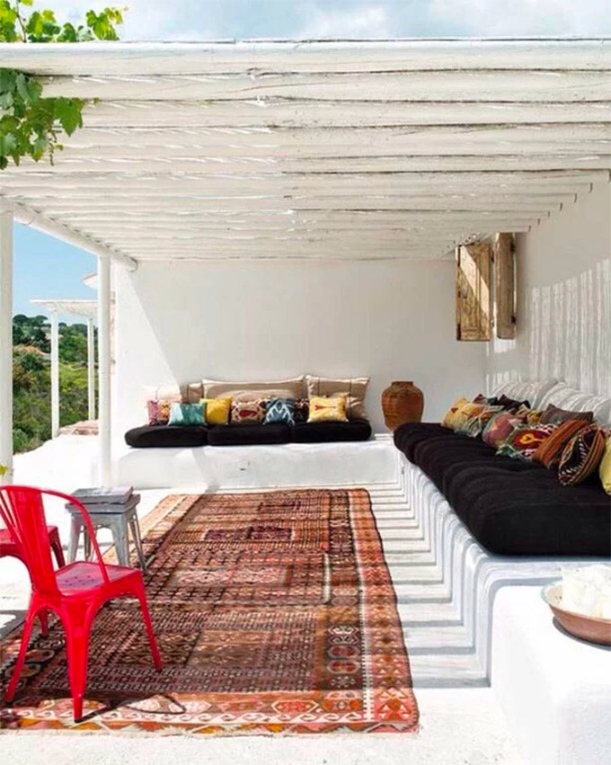 In de overdekte loungehoek van deze Ibiza tuin, is een kleurrijke bohemian sfeer gecreëerd met een groot vintage vloerkleed en gekleurde kussens op de banken.