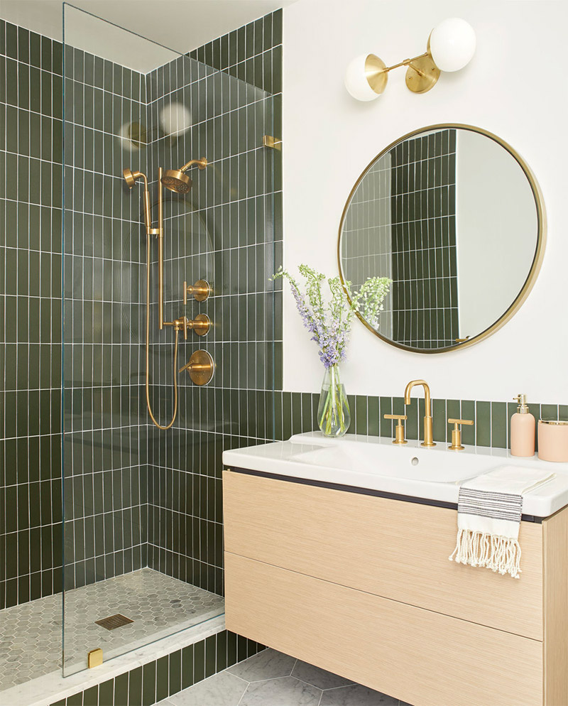 Deze badkamer met olijfgroene tegels is ontworpen door Clara Jung van Banner Day Interiors. De tegels staan prachtig bij de gouden kranen en de houten badkamermeubel.
