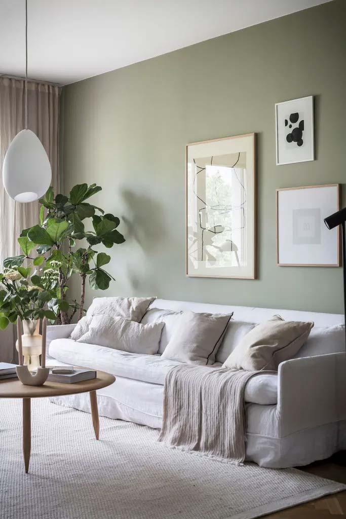 De sage green muren geven deze woonkamer een zachte kalmerende uitstraling, gecombineerd met de lichte bank, houten accenten en groene planten.