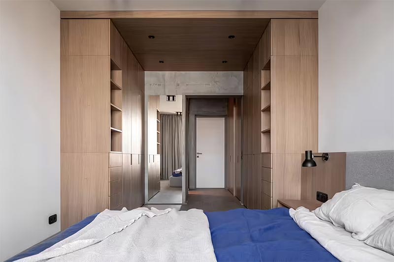Dit is de mooie slaapkamer van het Zarichnyy Apartment project, ontworpen door FILD, waar een soort inloopkast is gerealiseerd met inbouwkasten op maat aan twee zijden. Door ook het plafond te verlagen met een ombouw van hetzelfde eikenhout, is het een mooi intiem geheel geworden. | Fotografie: Andrey Bezuglov