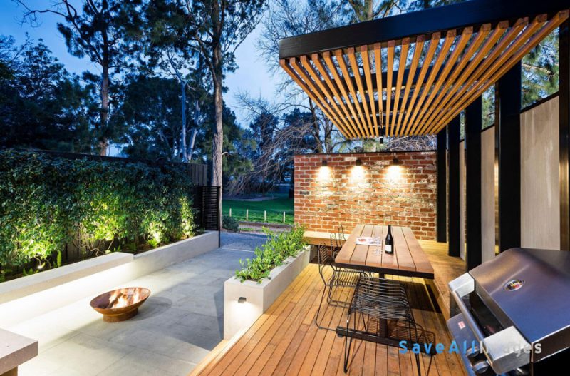 Deze moderne tuin combineert uplighting in de vorm van spotjes in de vlonders met downlighting in de vorm van moderne wandlampen aan de bakstenen schutting.
